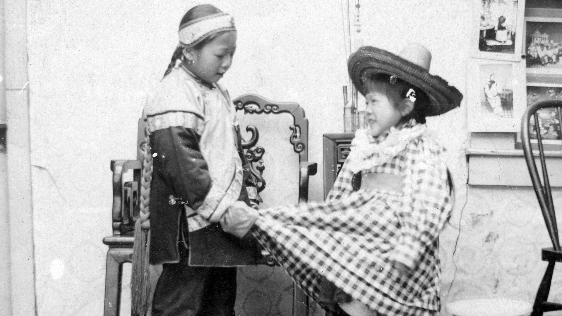 Black and white photograph depicting two children dressed in traditional Mexican attire, holding hands indoors with framed pictures on walls behind them. One child wears a sombrero and checkered poncho, while the other wears a headband and embroidered jacket, highlighting cultural dress and interaction.