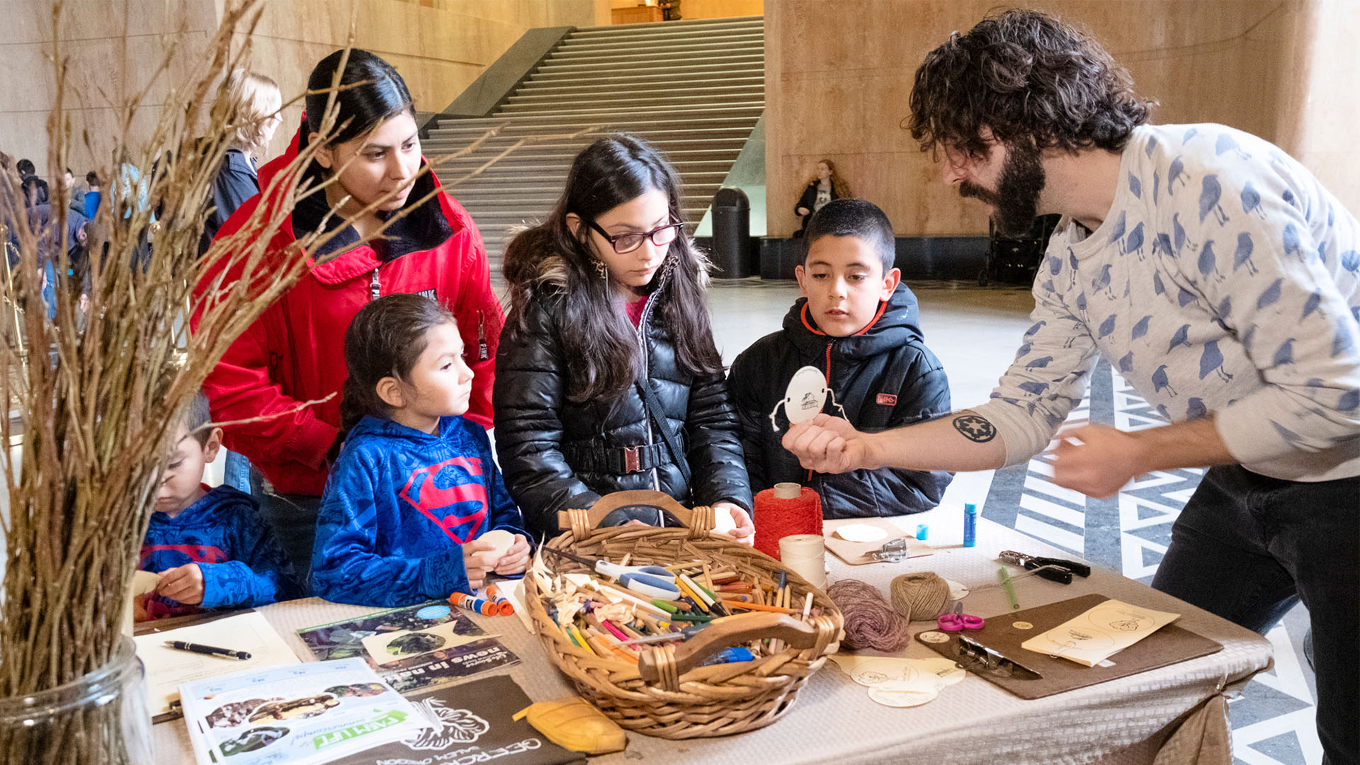 Photograph of a craft workshop where a man with curly hair and a beard demonstrates a small object to a group of children and adults gathered around a table. Table contains baskets with crafting supplies like yarn, scissors, and paper, set in a spacious indoor area with stairs and columns in the background.