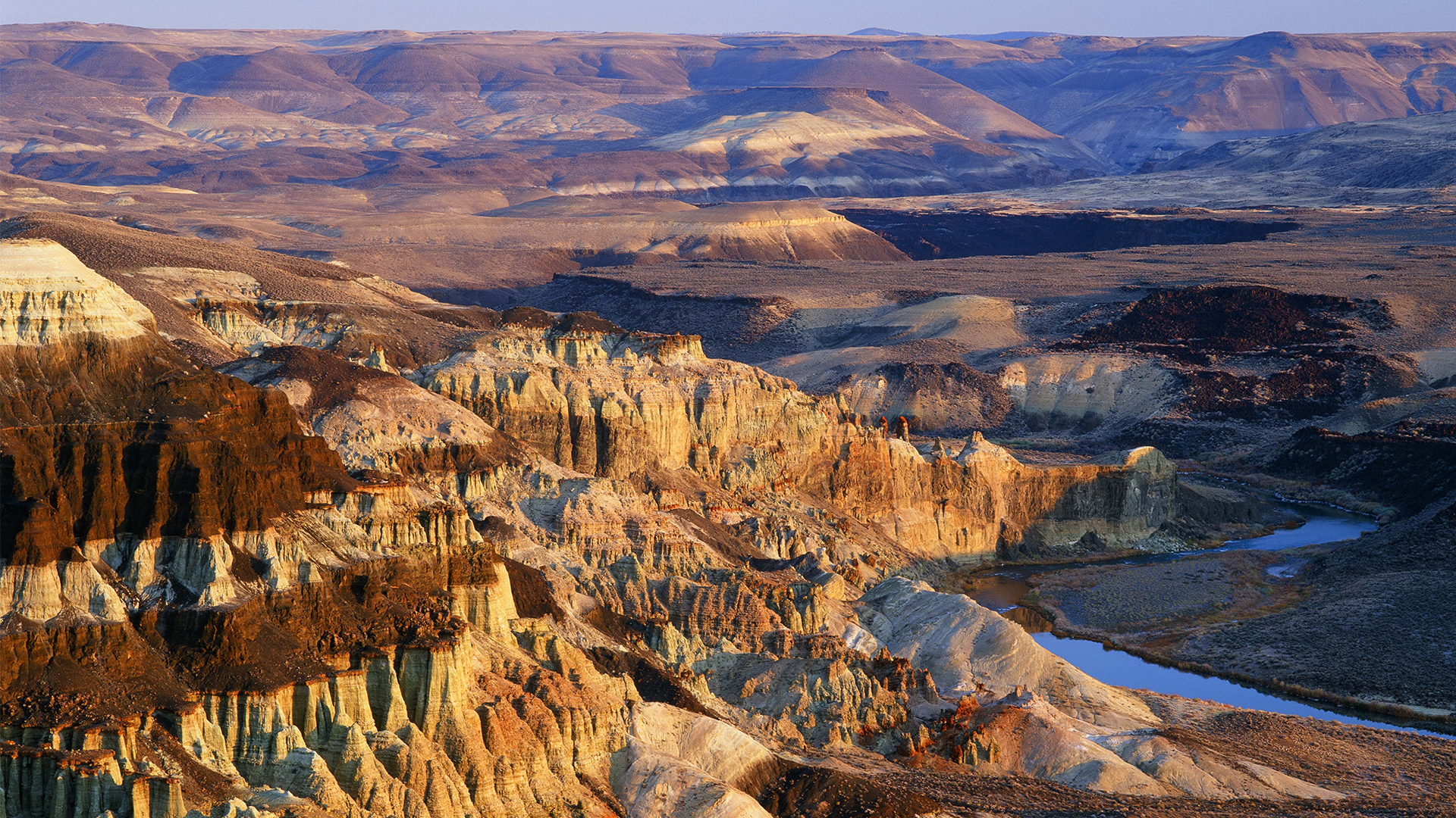 Photograph of a rugged canyon landscape with layered rock formations illuminated by warm sunlight, showcasing various shades of brown, beige, and orange. A winding river flows through the canyon, highlighting geological features and depth variations across the terrain.