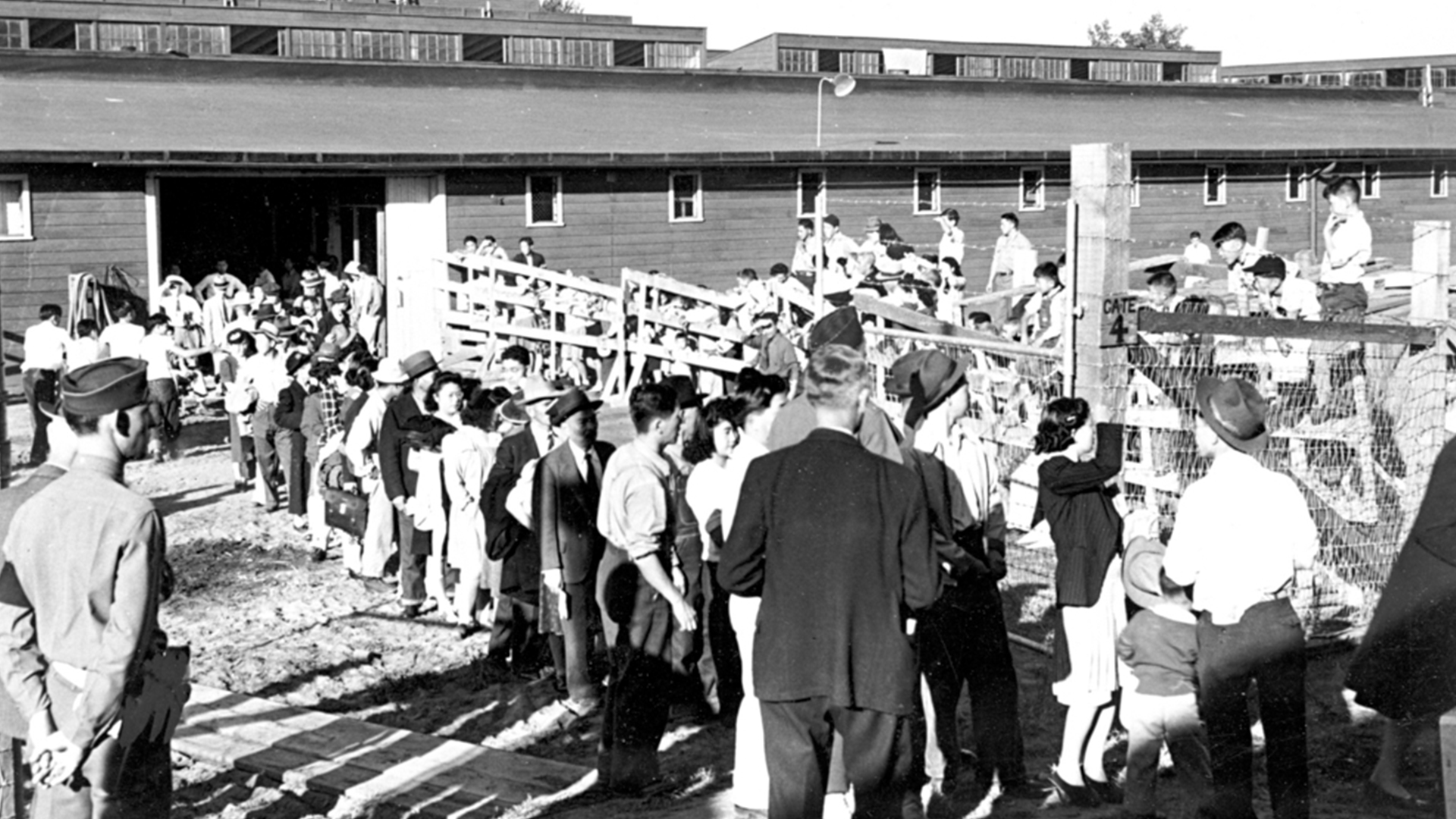 Black and white photograph showing a large group of people, including children and adults, lined up outside wooden barracks with barbed wire fencing. The scene depicts a historical setting related to wartime incarceration, highlighting the controlled and confined environment.