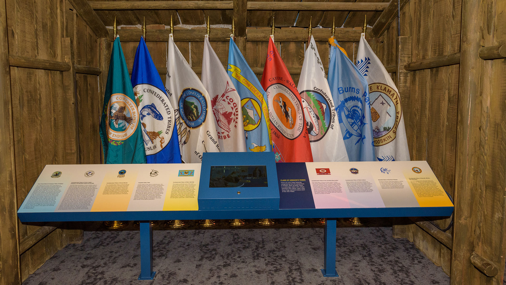 Photograph of a museum exhibit displaying nine colorful flags representing different U.S. military branches, arranged in a row behind an informational panel. The panel includes text and symbols with a central screen, set within a wooden structure that frames the display.