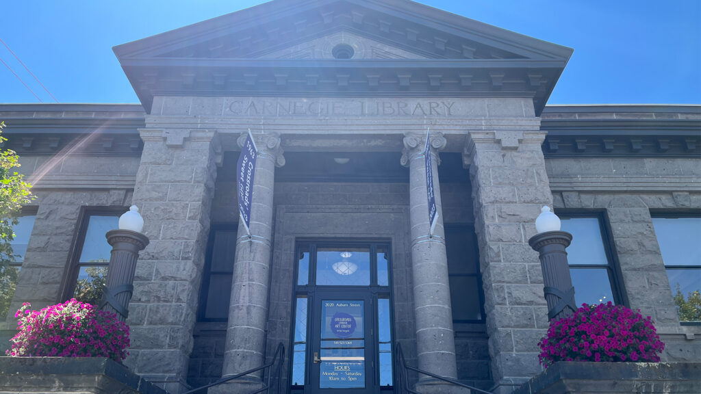 Photograph of a historic Carnegie Library building featuring a stone facade with four large columns and two decorative lamp posts topped with white globes. Bright purple flowers in planters flank the entrance, with banners hanging from the columns and a clear blue sky overhead.