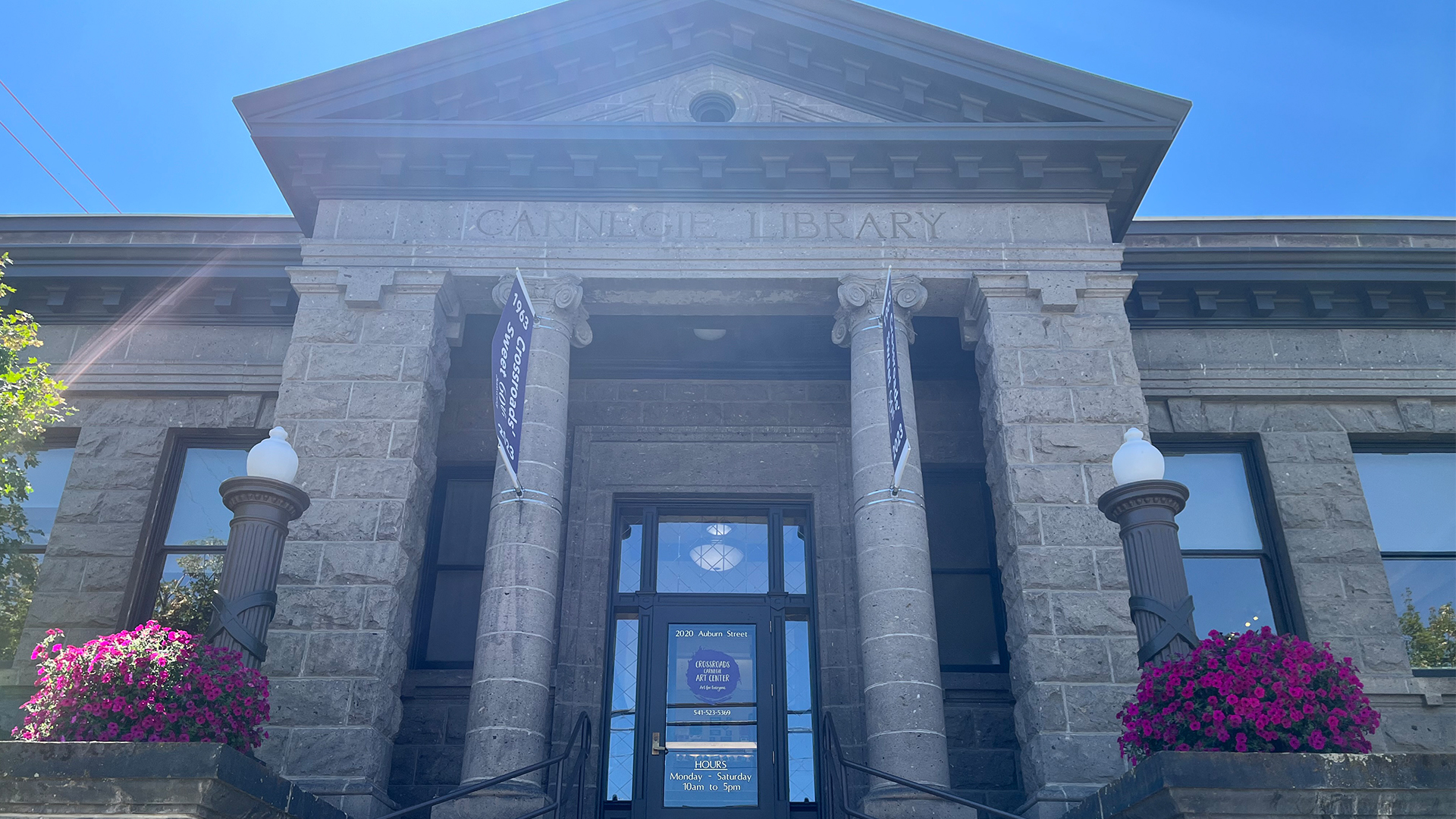 Photograph of a historic Carnegie Library building featuring a stone facade with four large columns and two decorative lamp posts topped with white globes. Bright purple flowers in planters flank the entrance, with banners hanging from the columns and a clear blue sky overhead.