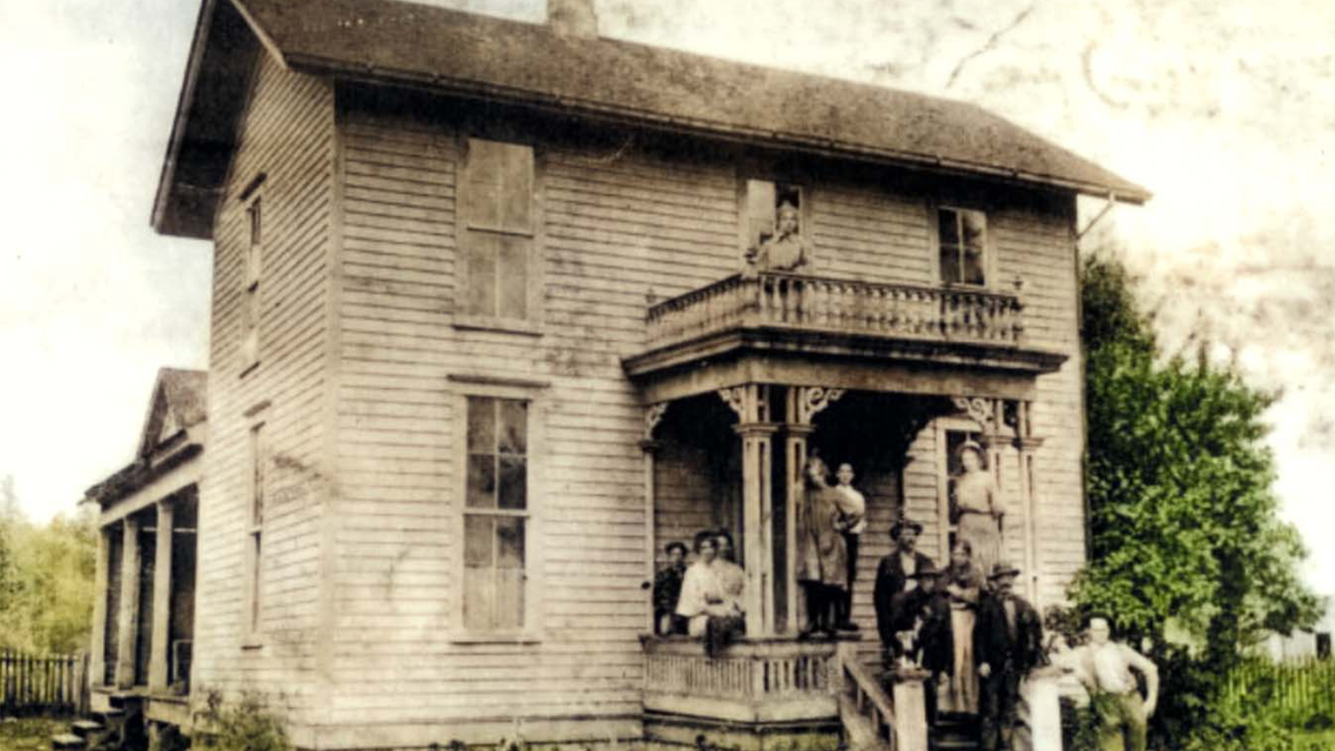 Photograph of an old two-story wooden house with a group of people posed on the front porch and balcony. The house features a covered porch with decorative columns and a railing on the balcony, set against a backdrop of trees and open sky.