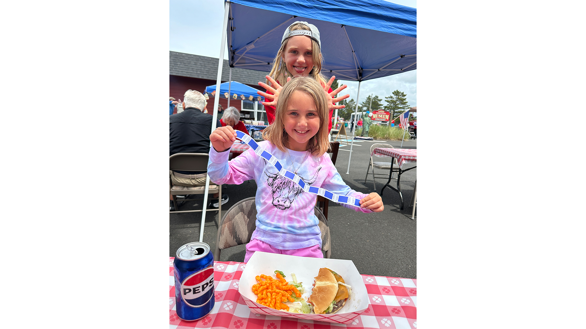 Photograph of two children at an outdoor event with picnic tables and tents, one child sitting and holding a strip of raffle tickets while the other stands behind making a playful hand gesture. The seated child wears a tie-dye shirt with a cow graphic, and a meal consisting of a burger, cheese curls, and a can of Pepsi is on a red and white checkered tablecloth in front of them.