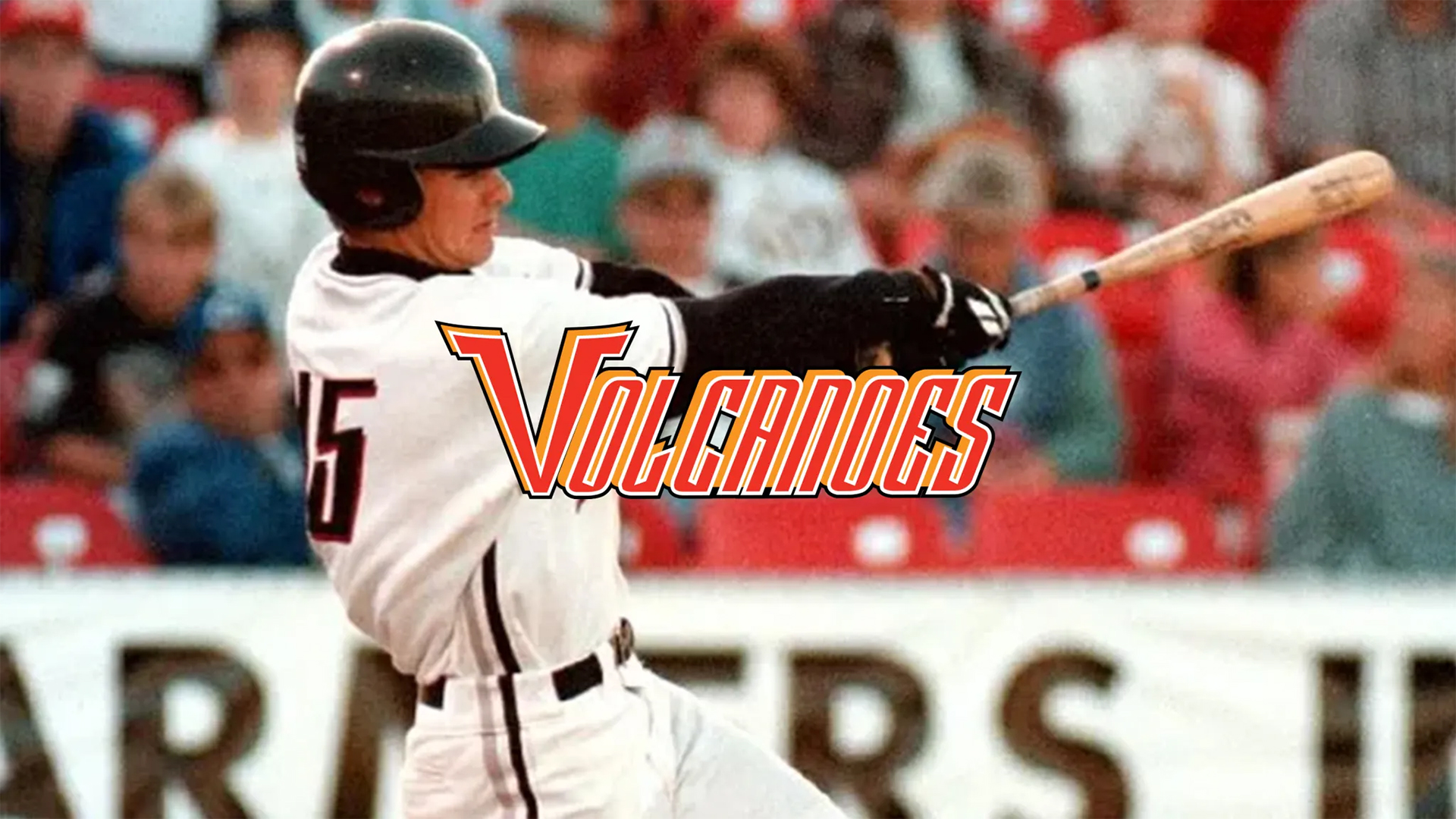 Photograph of a baseball player in white uniform swinging a bat during a game, with a crowd in the background. Bold orange and yellow "Volcanoes" text overlay is centered across the player.